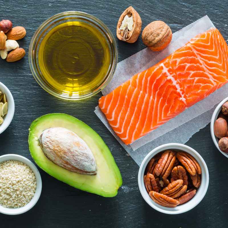 En la fotografía aparece diferentes alimentos ricos en grasas saludables: aceite de oliva, salmón, nueces, aguacate y avellanas.